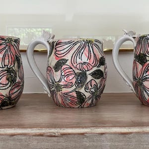 May include: Three ceramic mugs with white handles and floral designs. The mugs feature pink flowers with black outlines and green leaves on a cream background. Each mug has a small decorative element near the handle.