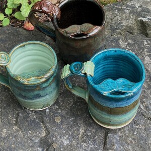 May include: Three handmade ceramic mugs featuring a moustache-shaped guard on the rim and a spiral detail on the handle. The mugs are finished in glossy glazes of dark brown, light green, and vibrant blue, each with a unique colour variation.