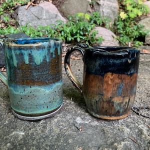 Può includere: Due tazze in ceramica con manici. La tazza a sinistra presenta uno smalto color ottanio e marrone con una consistenza maculata. La tazza a destra ha una parte superiore nera lucida che sfuma in una base marrone e blu, entrambe dall'aspetto rustico.