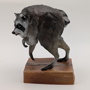 May include: A small figurine of a raccoon with gray and black fur, a white face mask, and a small pink tongue visible. The raccoon is depicted in a hunched, walking posture, mounted on a rectangular, light brown base with a wood grain texture.