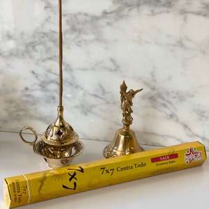 Könnte beinhalten: Eine Schachtel mit 7x7 Contra Todo Salbei-Räucherstäbchen mit 20 Stäbchen. Die Schachtel ist gelb mit einem Sonnendesign und dem Text "7x7 Contra Todo" in Schwarz. Die Räucherstäbchen befinden sich in einem Messing-Räucherofen mit Griff und einer kleinen Engelsfigur auf einer Messingglocke.