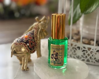 以下が含まれることがあります： 金色のキャップが付いた小さな緑色のガラス瓶で、「Feronomas en Aceite Mr. Money」とラベルが貼られています。白い長方形の石の上に置かれています。装飾的な毛布が付いた金の象の像が背景にあります。ボトルには緑色の液体が入っています。