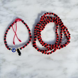 Puede incluir: Dos collares de cuentas y una pulsera. La joyería presenta cuentas rojas y negras alternas. La pulsera tiene un dije de ojo azul y un dije negro. La joyería se exhibe sobre una superficie de mármol blanco.