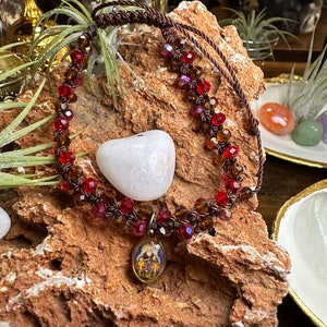Puede incluir: Un brazalete de cordón marrón con cuentas de vidrio rojas y marrones y un pequeño colgante con una imagen religiosa. El brazalete se muestra en una roca con una piedra blanca en forma de corazón.
