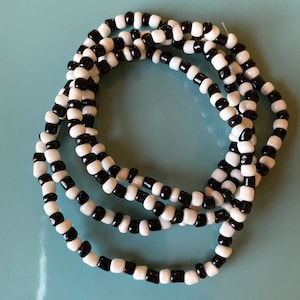 Puede incluir: Un collar de cuentas negras y blancas con múltiples hilos. Las cuentas son pequeñas y redondas, creando un diseño simple y elegante.
