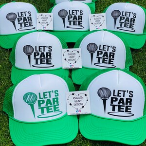 May include: A collection of green and white trucker hats with the text "LET'S PAR TEE" and a golf ball graphic. The hats have a mesh back and a curved brim. Small cards with the text "RUGGED HEART DESIGNS" are also visible.