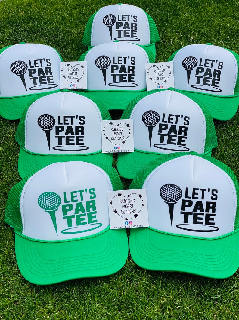 May include: A collection of green and white trucker hats with the text "LET'S PAR TEE" and a golf ball graphic. The hats have a mesh back and a curved brim. Small cards with the text "RUGGED HEART DESIGNS" are also visible.