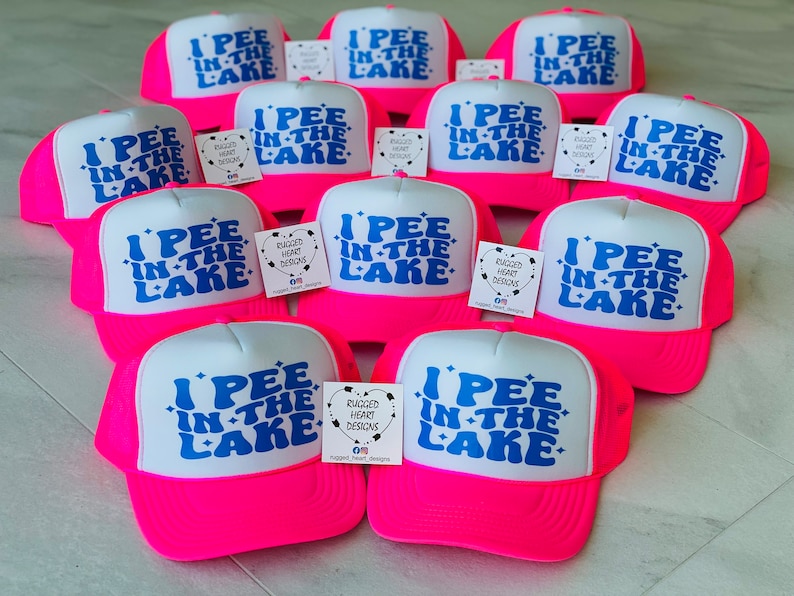 Pode incluir: Um grupo de bon&eacute;s de caminhoneiro rosa e branco com a frase "I PEE IN THE LAKE" impressa em azul na frente. Os bon&eacute;s t&ecirc;m uma parte de tr&aacute;s de malha e uma viseira branca. Cada bon&eacute; tem uma pequena etiqueta quadrada branca presa na frente com as palavras "RUGGED HEART DESIGNS" impressas nela.