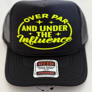 May include: Black trucker hat with a yellow graphic that reads "OVER PAR AND UNDER THE Influence". The hat has a mesh back and a curved brim. A tag on the front reads "OTTO WORLD FAMOUS FOAM FRONT TRUCKER".