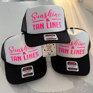 Puede incluir: Tres gorras de camionero con paneles frontales de espuma blanca y parte trasera de malla negra. Cada gorra presenta el texto "Sunshine & TAN LINES" impreso en rosa brillante. Tienen viseras negras y pegatinas de la marca OTTO.