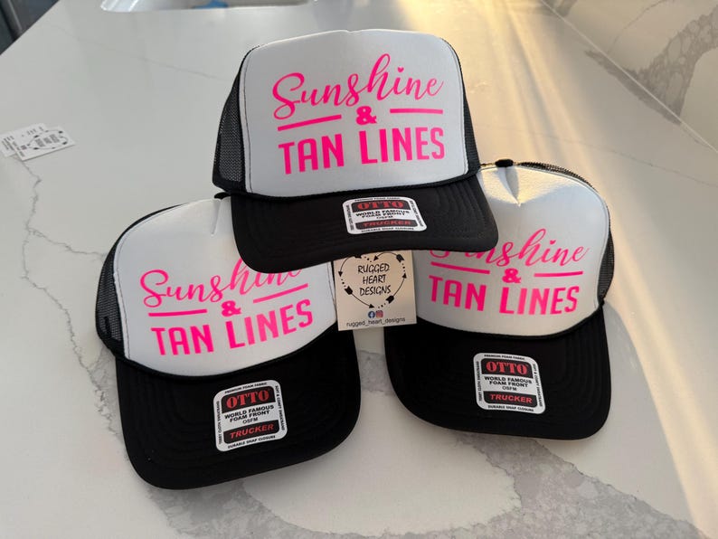 Puede incluir: Tres gorras de camionero con paneles frontales de espuma blanca y parte trasera de malla negra. Cada gorra presenta el texto "Sunshine & TAN LINES" impreso en rosa brillante. Tienen viseras negras y pegatinas de la marca OTTO.