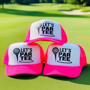 May include: Three neon pink and white trucker hats with the text "LET'S PAR TEE" and a golf ball graphic. The hats are displayed on a green golf course.