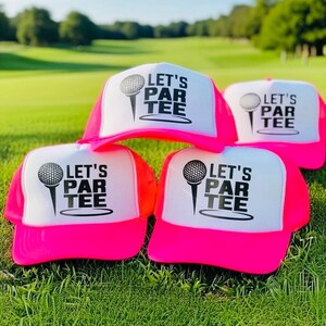 May include: A collection of neon pink and white trucker hats with the text "LET'S PAR TEE" and a golf ball graphic. The hats are displayed on a green grassy field, with a golf course in the background.
