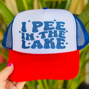 Pode incluir: Um chap&eacute;u de caminhoneiro branco, azul e vermelho com um design de glitter azul que diz "I PEE IN THE LAKE".