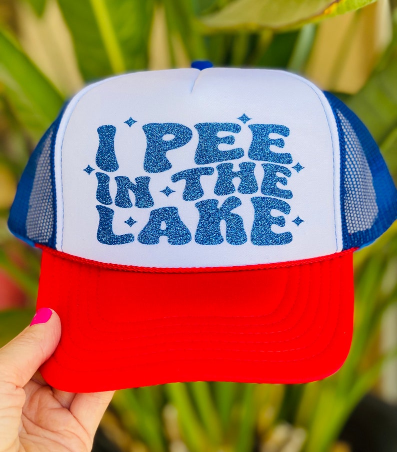 Pode incluir: Um chap&eacute;u de caminhoneiro branco, azul e vermelho com um design de glitter azul que diz "I PEE IN THE LAKE".