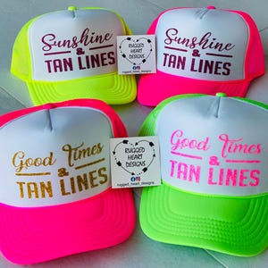 Puede incluir: Cuatro gorras de camionero con paneles frontales blancos y malla trasera de color ne&oacute;n. Dos gorras tienen el texto Sunshine & TAN LINES en granate brillante. Una tiene Good Times & TAN LINES en purpurina dorada y otra en rosa ne&oacute;n.