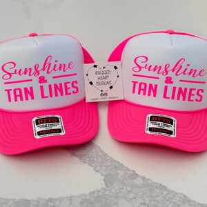 Puede incluir: Dos gorras de camionero de color rosa ne&oacute;n y blanco con el texto Sunshine & TAN LINES en letras rosas. Cada gorra tiene una pegatina negra y blanca de la marca Otto en la visera y una peque&ntilde;a etiqueta cuadrada con el texto Rugged Heart Designs.