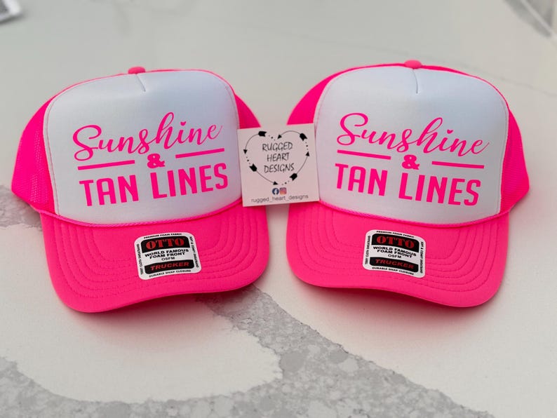 Puede incluir: Dos gorras de camionero de color rosa ne&oacute;n y blanco con el texto Sunshine & TAN LINES en letras rosas. Cada gorra tiene una pegatina negra y blanca de la marca Otto en la visera y una peque&ntilde;a etiqueta cuadrada con el texto Rugged Heart Designs.