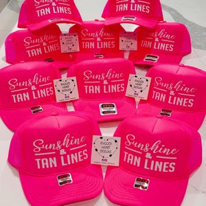 Puede incluir: Una colecci&oacute;n de gorras de camionero de color rosa ne&oacute;n brillante con el texto impreso en blanco Sunshine & TAN LINES. Cada gorra incluye un panel frontal estructurado, una parte trasera de malla y una etiqueta de marca en la visera.