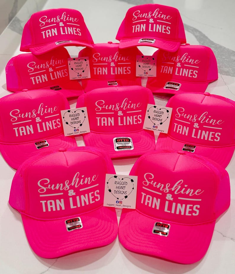 Puede incluir: Una colecci&oacute;n de gorras de camionero de color rosa ne&oacute;n brillante con el texto impreso en blanco Sunshine & TAN LINES. Cada gorra incluye un panel frontal estructurado, una parte trasera de malla y una etiqueta de marca en la visera.