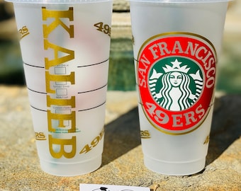 Könnte beinhalten: Zwei transparente Kunststoff-Kaltgetränkebecher mit Deckeln. Ein Becher hat den Namen "KALEB" in goldfarbenen Buchstaben, der andere zeigt ein San Francisco 49ers-Logo. Unten befindet sich eine kleine weiße Karte mit dem Text "RUGGED HEART".