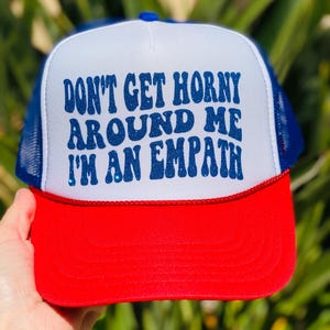 May include: A red, white, and blue trucker hat with the text "DON'T GET HORNY AROUND ME I'M AN EMPATH" in blue glitter lettering. The hat has a white front panel, a blue mesh back, and a red brim.