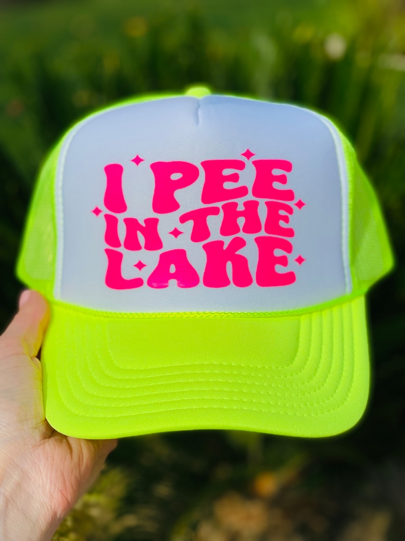 Pode incluir: Um chap&eacute;u de caminhoneiro amarelo neon e branco com um gr&aacute;fico rosa que diz "I PEE IN THE LAKE".