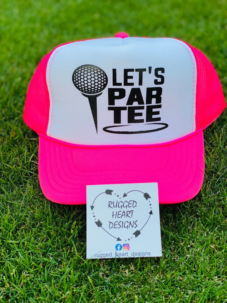 May include: A neon pink and white trucker hat with the words "LET'S PAR TEE" printed on the front, along with a golf ball and tee graphic. The hat is sitting on green grass. A small white card with the text "RUGGED HEART DESIGNS" is also visible.