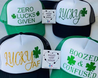 May include: Four trucker hats with different green and black designs. The hats have white mesh panels and feature the following text: "Zero Lucks Given", "Lucky", "Lucky AF", and "Boozed & Confused".