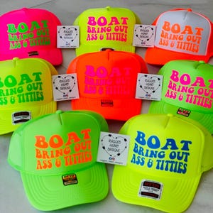 May include: A collection of neon trucker hats in various colors, including pink, orange, green, and yellow. Each hat features the phrase "BOAT BRING OUT ASS & TITTIES" in bold, contrasting colors. The hats have a mesh back and a curved brim.