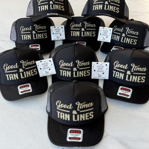 Puede incluir: Una colecci&oacute;n de gorras tipo camionero negras con parte trasera de malla y panel frontal de espuma. Cada gorra muestra el texto dorado Good Times & TAN LINES. Varias incluyen una etiqueta blanca con el texto RUGGED HEART DESIGNS.