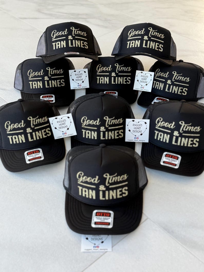 Puede incluir: Una colecci&oacute;n de gorras tipo camionero negras con parte trasera de malla y panel frontal de espuma. Cada gorra muestra el texto dorado Good Times & TAN LINES. Varias incluyen una etiqueta blanca con el texto RUGGED HEART DESIGNS.