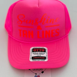 Puede incluir: Una gorra de camionero de color rosa ne&oacute;n brillante con un panel frontal de espuma que lleva impreso el texto naranja Sunshine & TAN LINES. Una pegatina rectangular blanca en la visera muestra OTTO WORLD FAMOUS FOAM FRONT OSFM TRUCKER DURABLE SNAP CLOSURE.