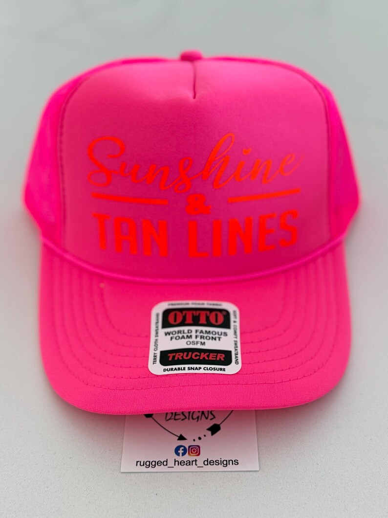 Puede incluir: Una gorra de camionero de color rosa ne&oacute;n brillante con un panel frontal de espuma que lleva impreso el texto naranja Sunshine & TAN LINES. Una pegatina rectangular blanca en la visera muestra OTTO WORLD FAMOUS FOAM FRONT OSFM TRUCKER DURABLE SNAP CLOSURE.