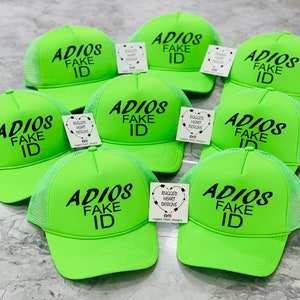 May include: A group of seven neon green trucker hats with black mesh backs. Each hat has the phrase "ADIOS FAKE ID" embroidered in black on the front. The hats are arranged in a semi-circle on a white surface.