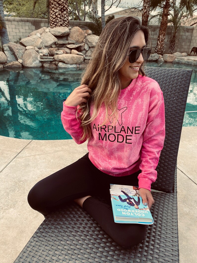 May include: A woman wearing a pink tie-dye sweatshirt with the text "AIRPLANE MODE" in black. She is sitting on a wicker chair by a pool, holding a blue book titled "Colton Underwood: First Time" in her hands.