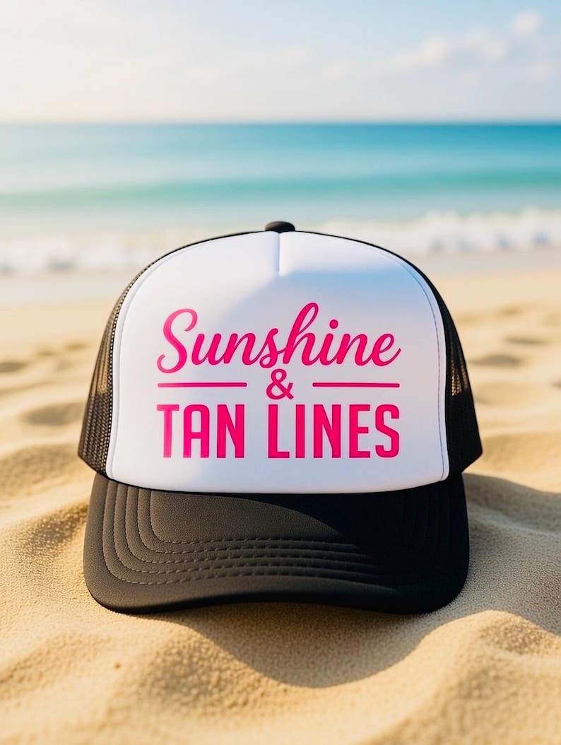 Puede incluir: Una gorra de camionero negra y blanca con un panel frontal blanco que presenta el texto en rosa intenso Sunshine & TAN LINES impreso en una mezcla de fuentes cursivas y de bloque. La gorra incluye una visera negra y paneles de malla negros.