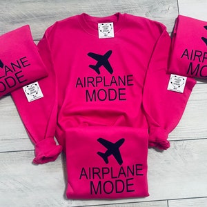 May include: Four pink t-shirts with the text "Airplane Mode" and a black airplane icon. The shirts are arranged in a pile on a light wood surface.