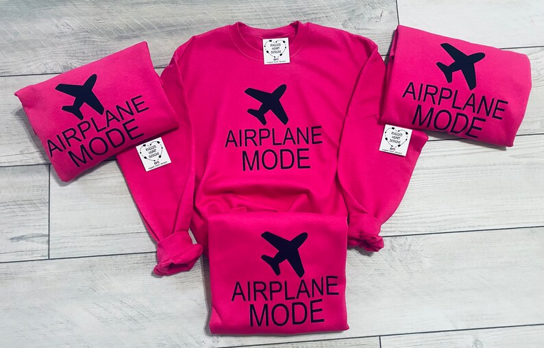 May include: Four pink t-shirts with the text "Airplane Mode" and a black airplane icon. The shirts are arranged in a pile on a light wood surface.
