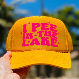 Pode incluir: Um bon&eacute; de caminhoneiro amarelo com um gr&aacute;fico rosa que diz "I PEE IN THE LAKE".