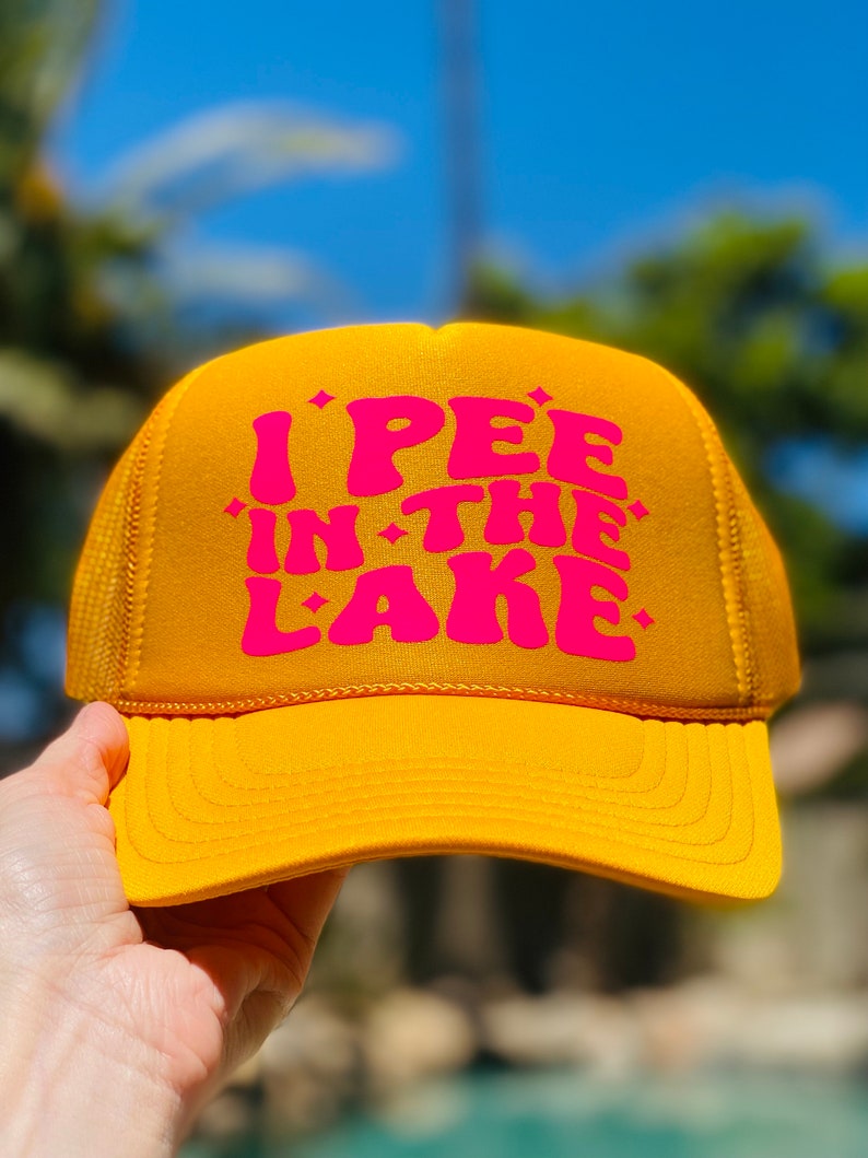 Pode incluir: Um bon&eacute; de caminhoneiro amarelo com um gr&aacute;fico rosa que diz "I PEE IN THE LAKE".