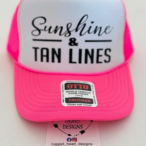 Puede incluir: Una gorra de camionero rosa ne&oacute;n y blanca con el texto negro Sunshine & TAN LINES en el panel frontal de espuma. Una pegatina rectangular en la visera dice OTTO WORLD FAMOUS FOAM FRONT TRUCKER. Una etiqueta blanca muestra HEART DESIGNS.