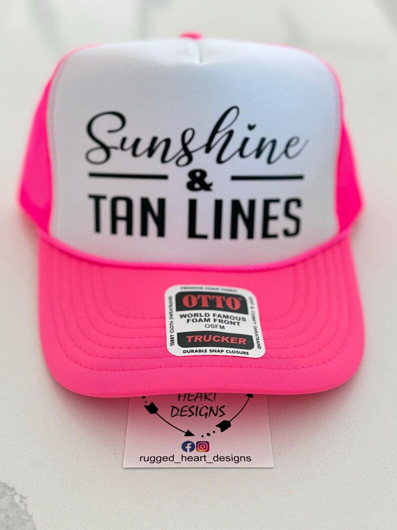 Puede incluir: Una gorra de camionero rosa ne&oacute;n y blanca con el texto negro Sunshine & TAN LINES en el panel frontal de espuma. Una pegatina rectangular en la visera dice OTTO WORLD FAMOUS FOAM FRONT TRUCKER. Una etiqueta blanca muestra HEART DESIGNS.