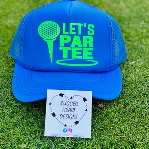 May include: A blue trucker hat with a neon green golf ball and tee graphic, along with the words "LET'S PAR TEE." The hat has a mesh back and a curved brim. A small card with the text "RUGGED HEART DESIGNS" is also visible.