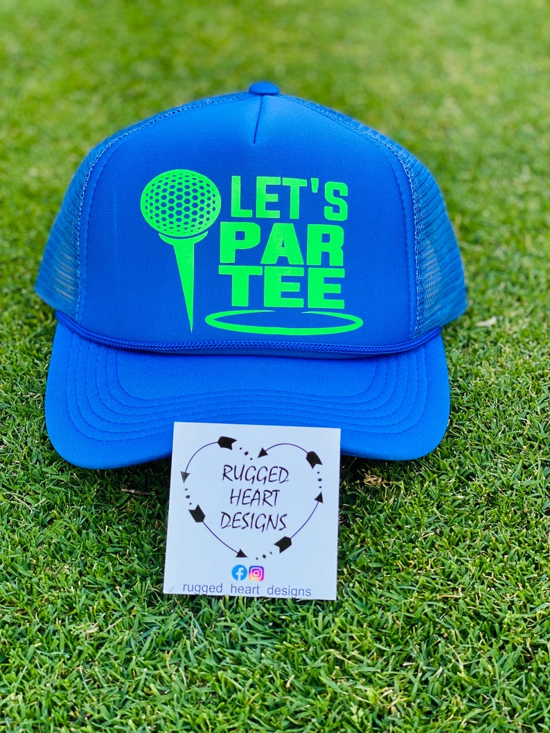 May include: A blue trucker hat with a neon green golf ball and tee graphic, along with the words "LET'S PAR TEE." The hat has a mesh back and a curved brim. A small card with the text "RUGGED HEART DESIGNS" is also visible.