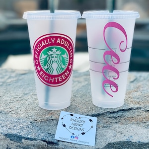 May include: Two clear plastic cups with pink and green designs. The cup on the left has a green Starbucks logo with the text "Officially Adulting Eighteen". The cup on the right has a pink cursive letter "C".