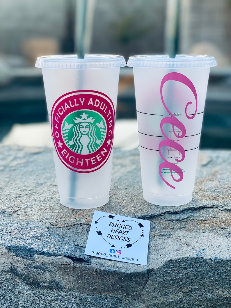 May include: Two clear plastic cups with pink and green designs. The cup on the left has a green Starbucks logo with the text "Officially Adulting Eighteen". The cup on the right has a pink cursive letter "C".