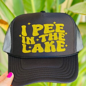 Pode incluir: Bon&eacute; de caminhoneiro preto com um design de glitter dourado que diz "I PEE IN THE LAKE".