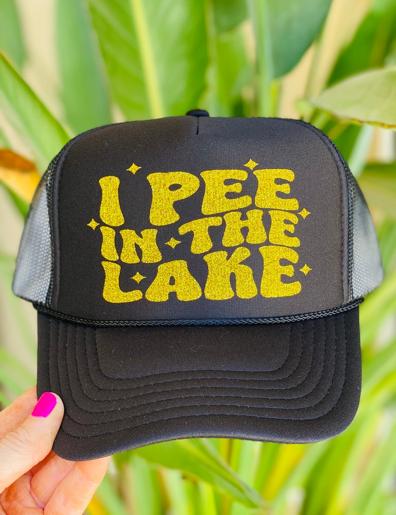 Pode incluir: Bon&eacute; de caminhoneiro preto com um design de glitter dourado que diz "I PEE IN THE LAKE".