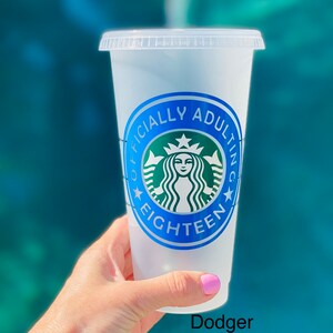 May include: A clear plastic reusable cup with a blue and green sticker that says "Officially Adulting Eighteen" with a stylized green Starbucks logo. The cup is being held by a hand with pink nail polish.